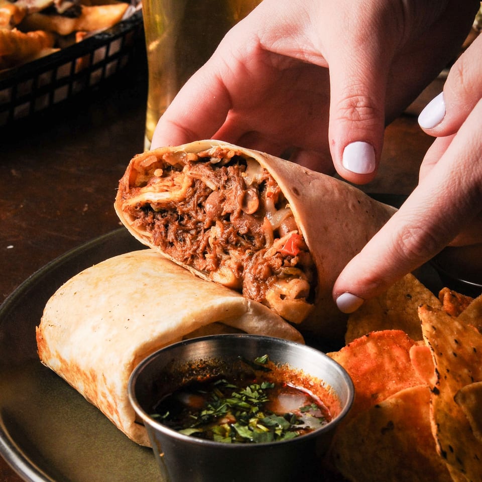 Hand serves a stuffed shawarma wrap alongside pita bread, dipping sauce, and a tall drink.