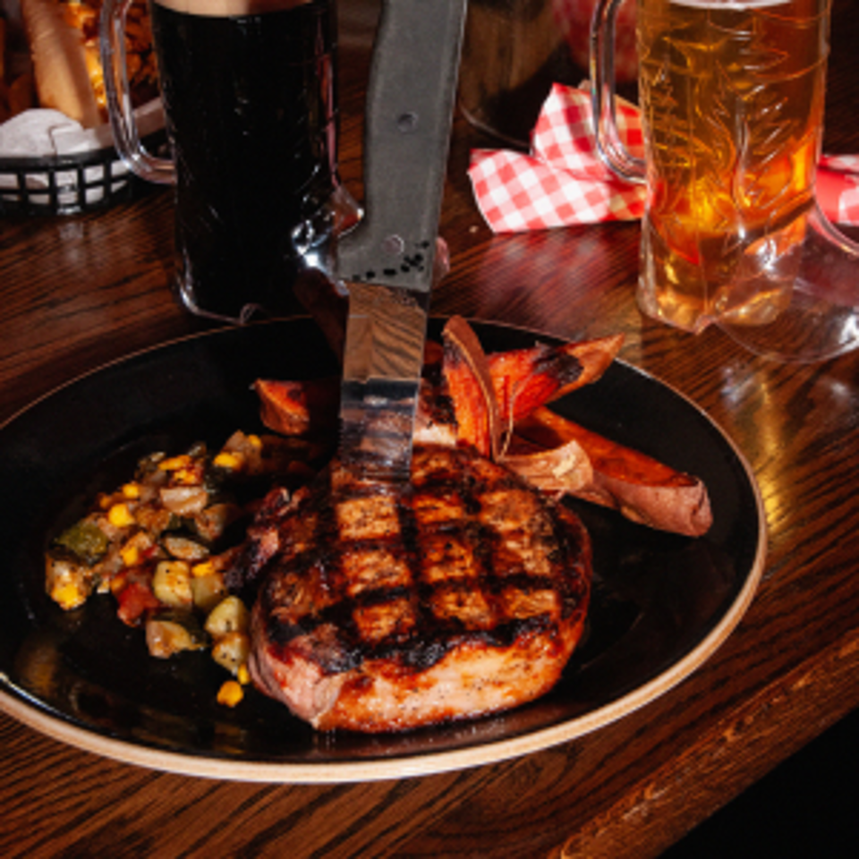 Grilled steak with crosshatch marks, roasted vegetables, and fries on a black plate beside beer glasses.