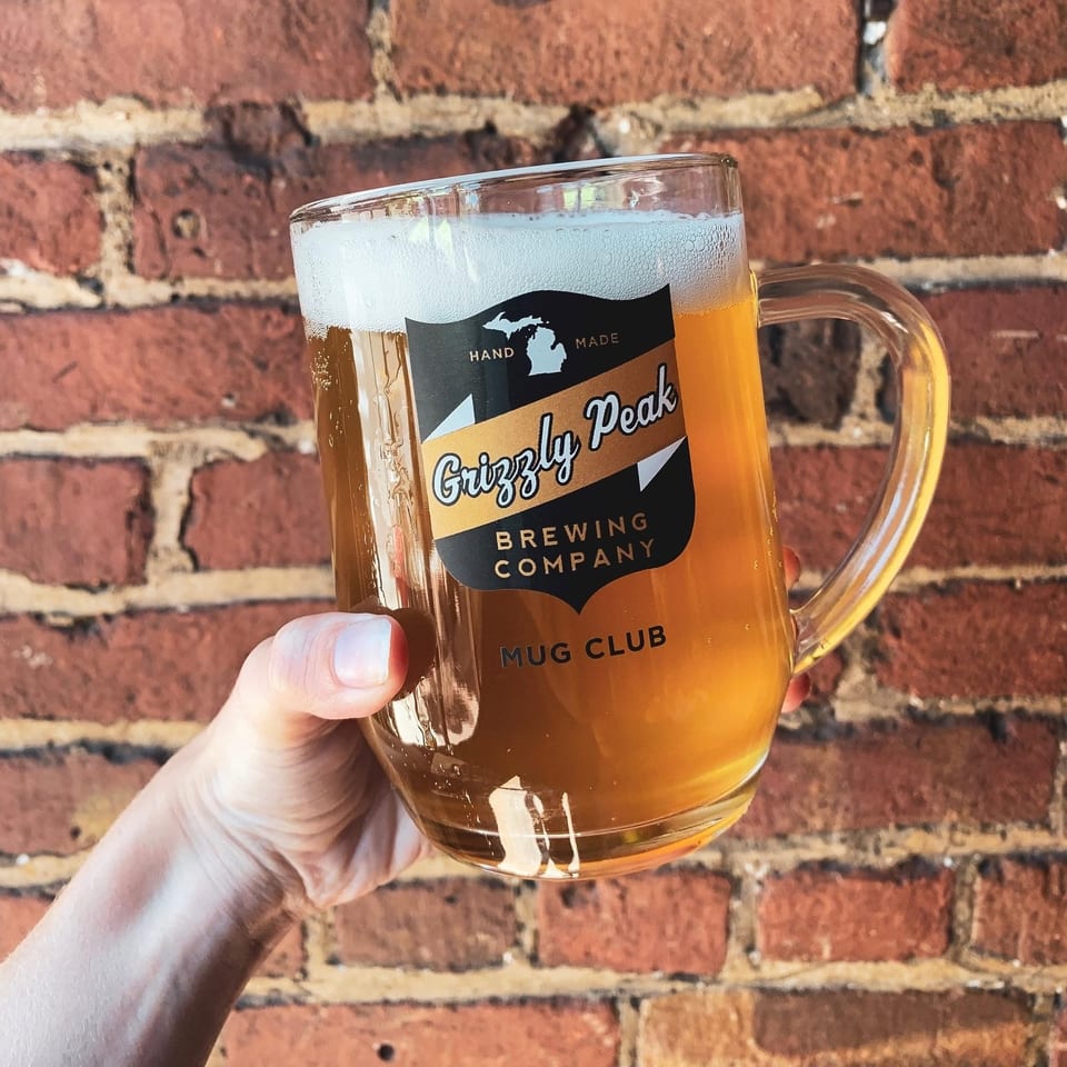 A hand holds a foamy amber Grizzly Peak Brewing Company beer mug against a red brick wall.