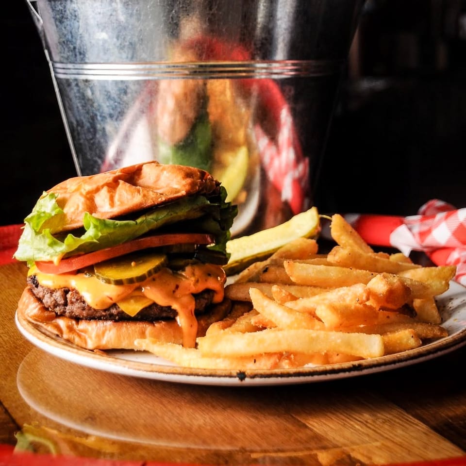 Cheeseburger topped with lettuce, tomato, pickles, and sauce served with fries and a pickle on a plate.