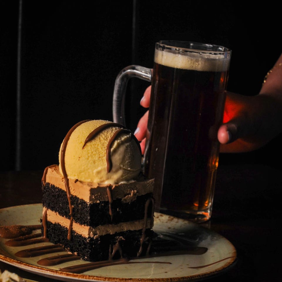 Chocolate cake with ice cream and chocolate drizzle beside a frothy dark beer, held by a diner in a dimly lit restaurant.